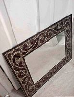 Rectangular mosaic framed mirror leaning against a white door, showing detailed mosaic tile border with swirl pattern in earth tones