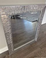 Silver textured rectangular mirror leaning against a light-colored wall on a wooden floor.