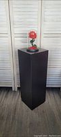 Black rectangular display pedestal approximately 20x20 inches with a decorative rose inside a glass dome placed on top for scale and presentation. Background white wood panel.