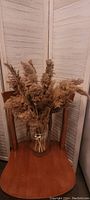 The glass vase filled with dried pampas grass placed on a wooden chair with a folding white shutter screen backdrop.