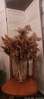 Glass vase filled with dried pampas grass set on a wooden table in front of a white wooden folding screen.