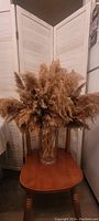 Image shows a clear glass vase with a wide mouth and pampas grass arranged inside it, viewed against a white folding screen and sitting on a wooden chair.