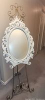White oval mirror with ornate floral frame and metal stand with scrollwork