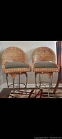 Pair of wicker and iron stools with cushions shown side by side on rug indoors near window and wooden table.