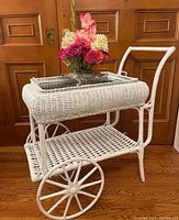Full view of the white wicker bar/tea cart showing front wheels, handle, and a vase of flowers on top.