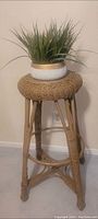 Photo showing a tall wicker high stool with a woven seat and wooden legs, featuring a potted artificial plant on top (plant not included in lot)