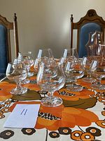 11 clear glass brandy snifters arranged on a floral tablecloth with orange and brown colors, showing their rounded bowl and short stem shape.
