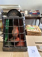 Front angle showing metal lantern with protective wire grid, glass panel, and central glass chimney on metal base. Nautical ship wheel motif at top.