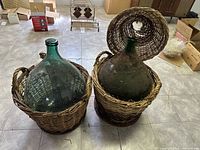 Two large green glass demijohns sitting in woven wicker baskets, one showing the removable basket lid.