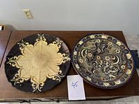 Photo of two art plates side by side on table, showing one with brown and beige raised symmetrical design, the other with dark blue and white bird and flower patterns.