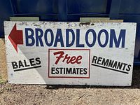 Full view of the large white rectangular wooden sign with blue, black and red painted letters showing 'BROADLOOM', directional arrow, and advertising text.