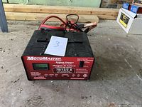 Front view of the black battery charger showing red faceplate with model name MotoMaster, digital display screen, and attached cables with clamps.