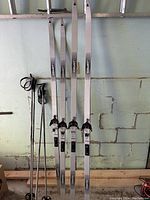 Two pairs of cross country skis standing upright against a wall with poles leaning beside them. The white and gray pair is on the left and the blue-gray pair is on the right.