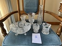 Full collection of six glass and crystal decorative pieces on blue fabric chair: a large footed bowl, two small vases, a covered dish, and two candle holders.