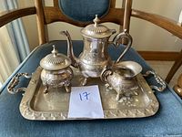 Photo of the brass tea set on a wooden chair, showing teapot, sugar bowl, creamer and tray with detailed handle and embossed patterns from a front angle.