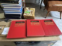 Three red hardcover Popular Mechanics Do-It-Yourself Encyclopedia books stacked side by side on a cardboard box, showing front covers clearly.