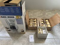 Photo showing the boxed Black & Decker coffee maker beside three boxes of new decorated coffee mugs on a tiled floor.
