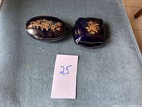 Two Italian porcelain trinket dishes side by side on a blue fabric. Both have cobalt blue glaze with gold floral decoration on the lids.