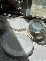 Stack of white ceramic plates and platters with textured rim design and gold trim, clear glass plates, and patterned ceramic plates stacked together near a window.