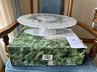 Clear glass cake platter displayed on original box with green foliage design and branding label visible.