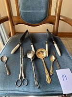 Seven utensils arranged on a blue chair, including spoons, knives with white handles, tongs and ladles.