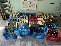 Wide shot of multiple blue and red plastic crates containing vintage glass pop bottles plus cardboard carriers on concrete floor.
