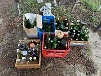 Overall view of the lot showing multiple crates and boxes filled with green and clear glass bottles outdoors on dirt ground.