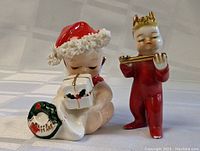 Two vintage ceramic Christmas figures on a white cloth background. One is a child holding a gift and stocking, other playing a flute.