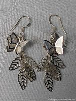 Pair of sterling silver earrings with butterfly and detailed leaf charms hanging from hooks shown against a grey fabric background.