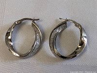 Pair of sterling silver hoop earrings resting on a light textured fabric background. Both earrings visible showing front and side views, highlighting the encrusted sparkling stones on the inside section of each hoop and polished outer surface.