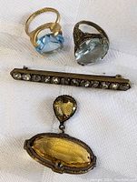 Four pieces of vintage costume jewelry: two light blue stone rings, a rhinestone bar brooch, and a yellow two-stone pendant on white fabric