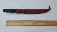 Photo showing the closed fillet knife with attached brown leather sheath and a ruler for size reference. The knife's handle is black with brass-colored accents.