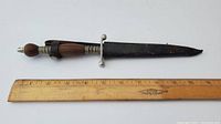 Dagger in sheath with ruler for size reference, showing overall condition and length about 9 inches including handle and sheath