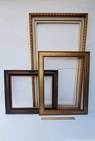 Photo showing three decorative picture frames leaning against a wall: a large gold-tone, a medium gold-tone, and a smaller dark brown frame.