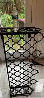 Full view of antique wrought iron fireplace cover standing upright with a water bottle on top for scale, showing frame and cutout pattern.