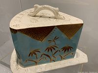 Side angle of the cheese dome showing turquoise ceramic with gold bamboo and geometric designs, and the cream-colored embossed base plate