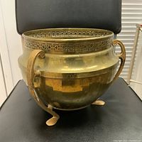 Front side view of the brass planter showing one handle, side foot, and the ornate pierced rim detail.
