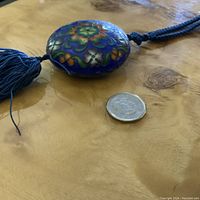 Close-up view of the round cloisonné pendant showing colorful floral enamel work with blue tassel and coin for size comparison.