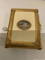 Rectangular ornate gold-tone music box with ivory panel lid and oval signed portrait in center, quarter placed for scale.