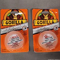 Photo showing two sealed packs of Gorilla Double-Sided Clear Mounting Tape, each roll 1 inch by 60 inches, in orange Gorilla packaging.