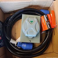 Power cord coiled inside a box with one grey inlet receptacle box on top, blue 4-prong male plug visible.