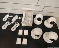 Four white indoor 360 security cameras arranged in a square, with four USB cables and four power adapters. Cameras have a spherical design with black lens and silver detailing.