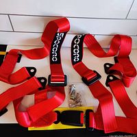 Full view of the red 4 point racing harness with attached fastening hardware and yellow buckle strap, laid out on white wooden floor.