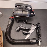 Complete set of black electric blower with attached hose, four nozzle attachments and removal tool laid out on black surface.