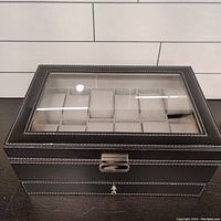 Closed view of black faux leather box with white stitching and glass top showing 12 watch compartments.