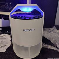 Front view of Katchy insect trap powered on with visible blue UV light and fan blades visible in black inside device.