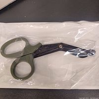 Top view of military trauma scissors in sealed plastic packaging, showing olive handles and black angled safety blades.