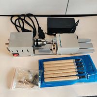 Full view of the mini lathe with power cable, chuck, tailstock, and 6 chisels in blue packaging along with small accessory tools in plastic bag