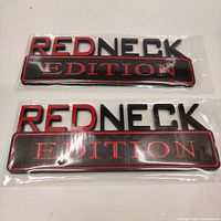 Two black and red 'REDNECK EDITION' vehicle emblems in sealed clear plastic packaging, one stacked above the other on a white surface.