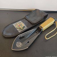 Full view of black steel survival shovel blade with serrated edge and paracord wrapped handle shown with nylon carrying pouch